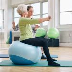 Eine ältere Frau hält unter Anleitung der Kursleitung beide Arme mit einem Gewicht ausgestreckt nach vorneach vorne 