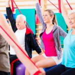 ältere Frau arbeitet unter Anleitung der Kurstrainerin mit einem Terraband schräg hinter dem Rücken